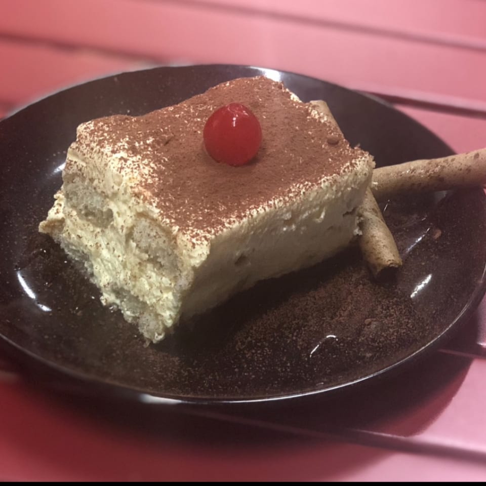 A slice of tiramisu with cocoa powder and a cherry on top, served on a dark plate.
