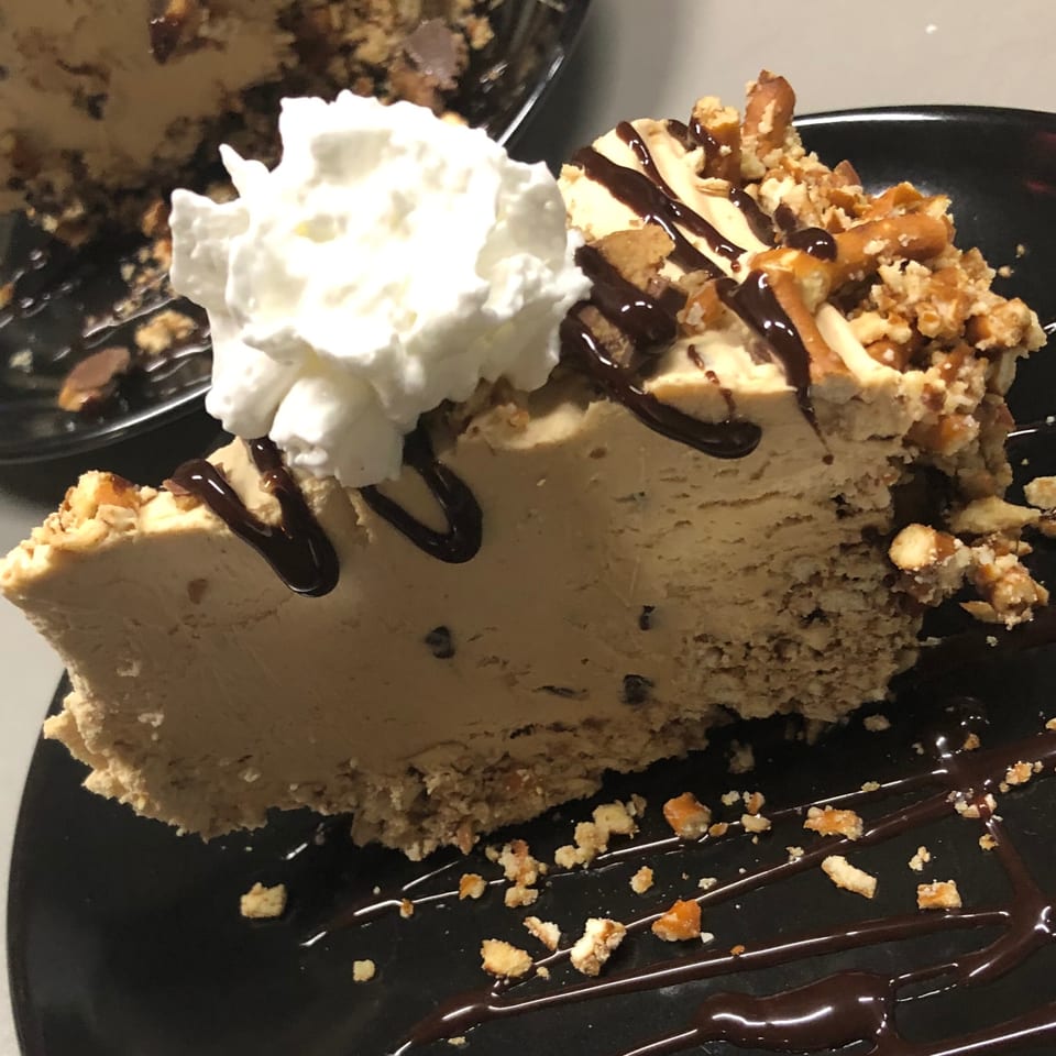 A slice of peanut butter pie with whipped cream, chocolate drizzle, and pretzel crumbles.