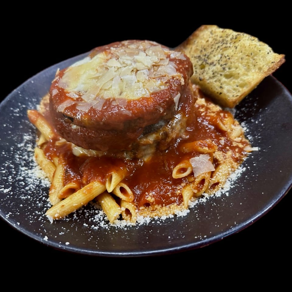 Meatball parmesan with penne pasta, red sauce, and garlic bread on a black plate.