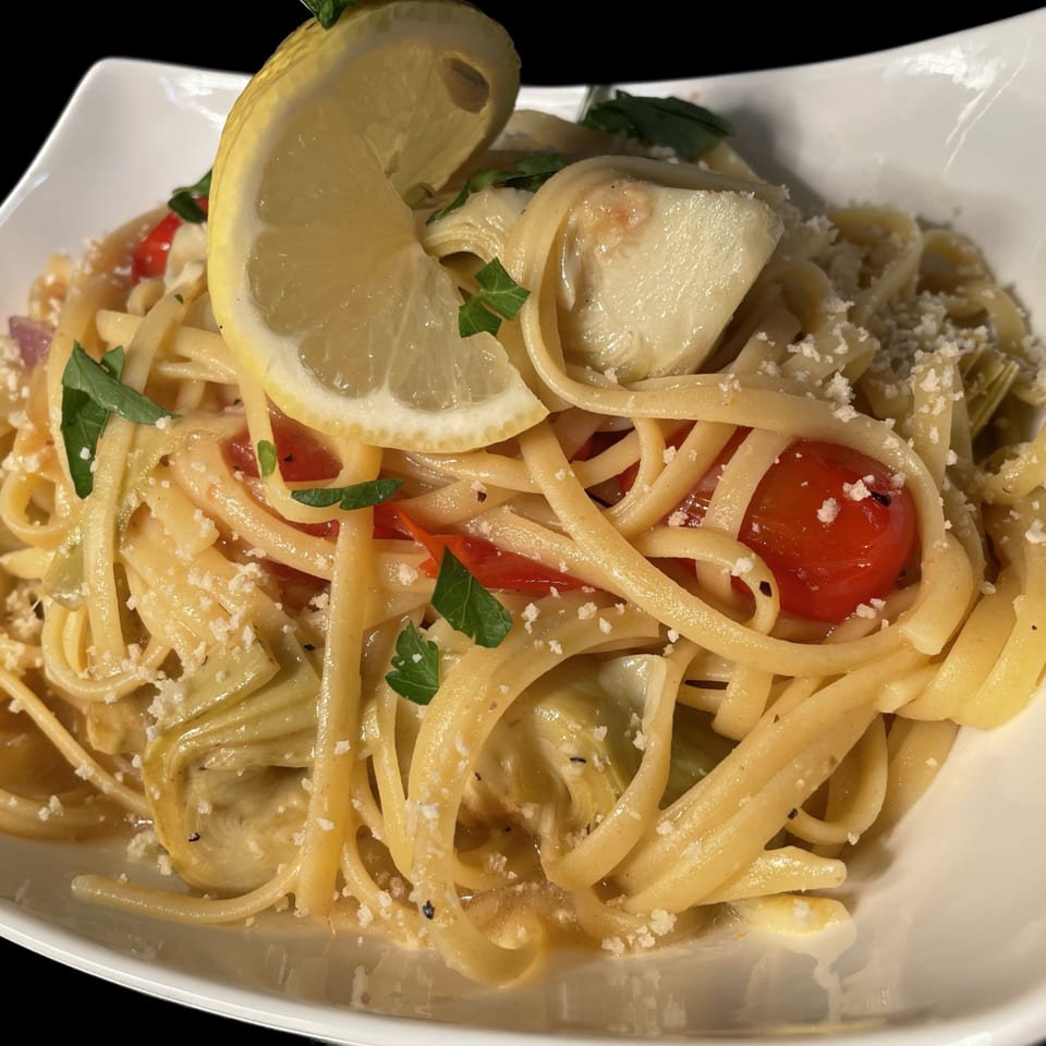 Linguine pasta with cherry tomatoes, artichoke hearts, parmesan, parsley, and a lemon wedge in a white bowl.