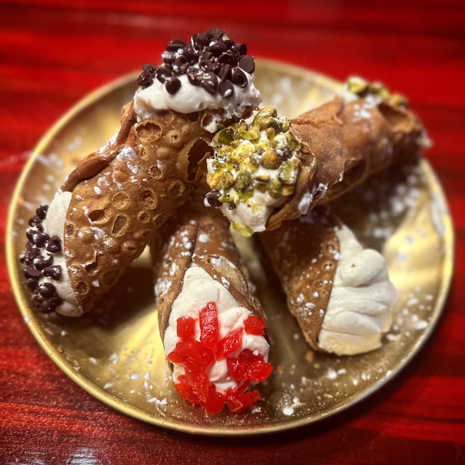 Four cannoli on a gold plate, two with chocolate chips, one with pistachios, and one with candied cherries.