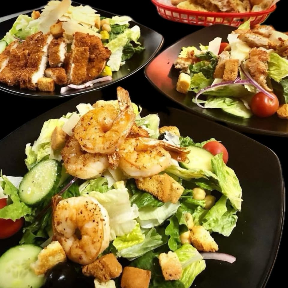 Three Caesar salads with chicken, shrimp, and croutons on black plates, with a bread basket.