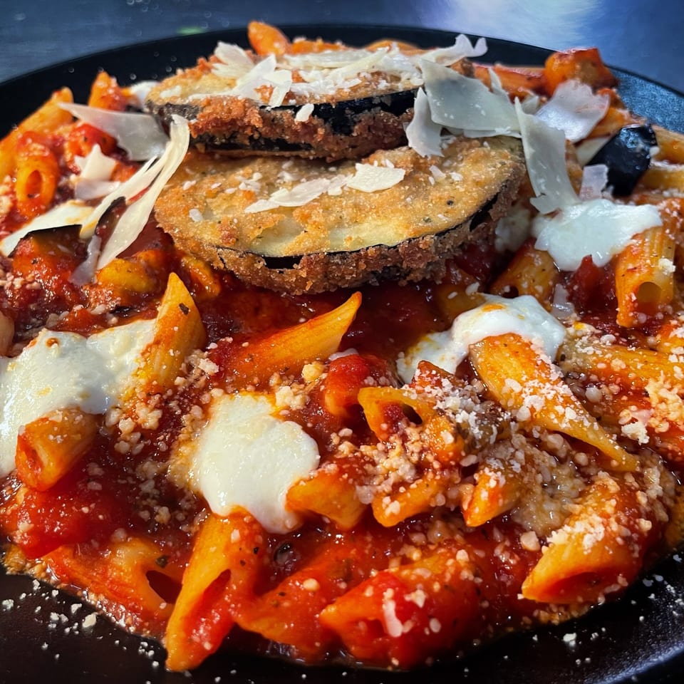 Penne pasta with red sauce, melted mozzarella, parmesan, and fried eggplant slices on a black plate.